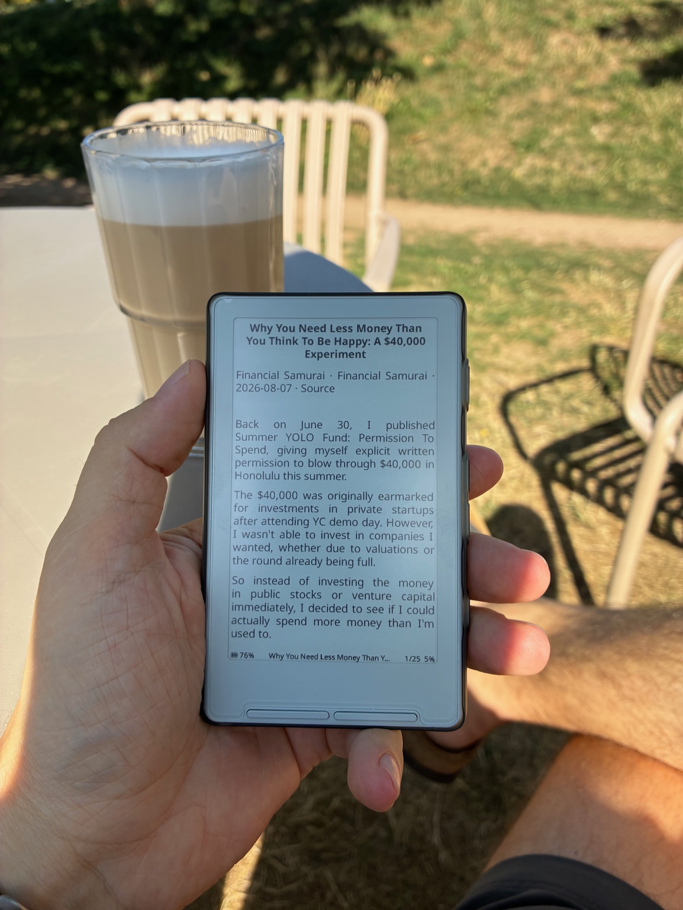 Рука держит небольшой e-ink ридер X4 на улице, на экране виден блог-пост. На заднем плане в солнечных лучах — латте и металлический стул кафе.