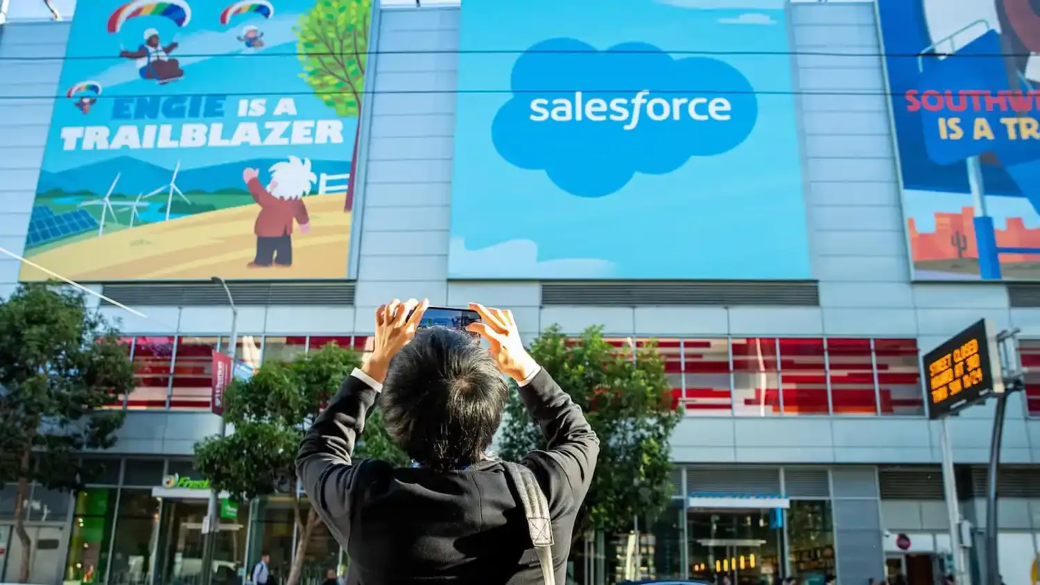 Кто-то фотографирует снаружи офисного здания Salesforce. На биллборде нарисованные маскоты и надпись 'Engie is a Trailblazer'
