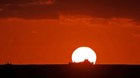 Getty Images The Sun sets over an ocean. The sky is dark red and the silhouette of a ship sailing across the ocean in front of the Sun.