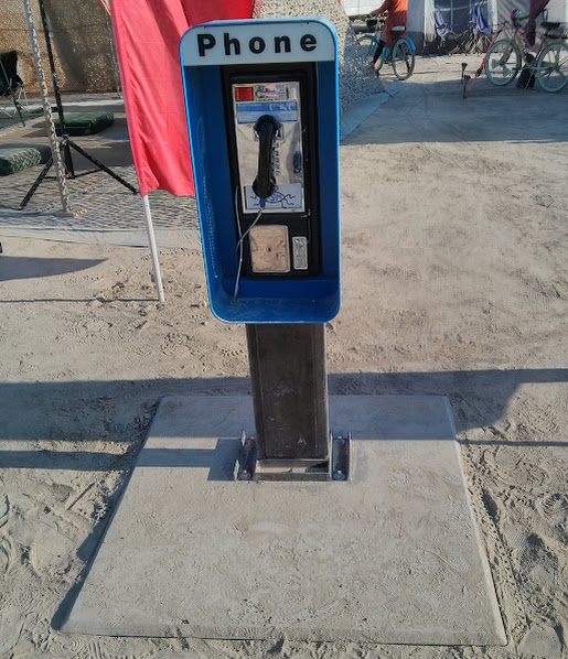 Будка Playa Phone на плайе: синий таксофон на чёрном столбе с вывеской PHONE, на фоне лагерного тента и велосипедов.