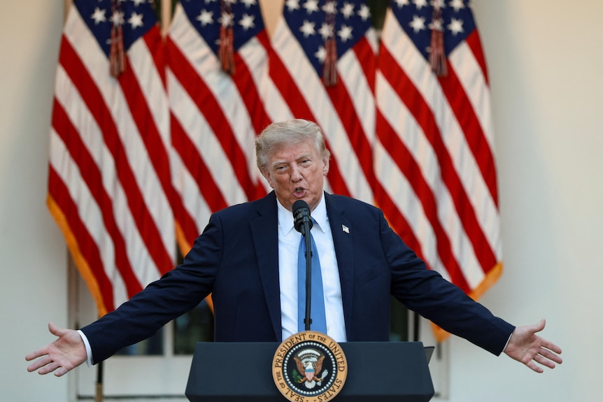 Дональд Трамп в чёрном костюме, белой рубашке и синем галстуке выступает у микрофона с расставленными руками перед флагами США.